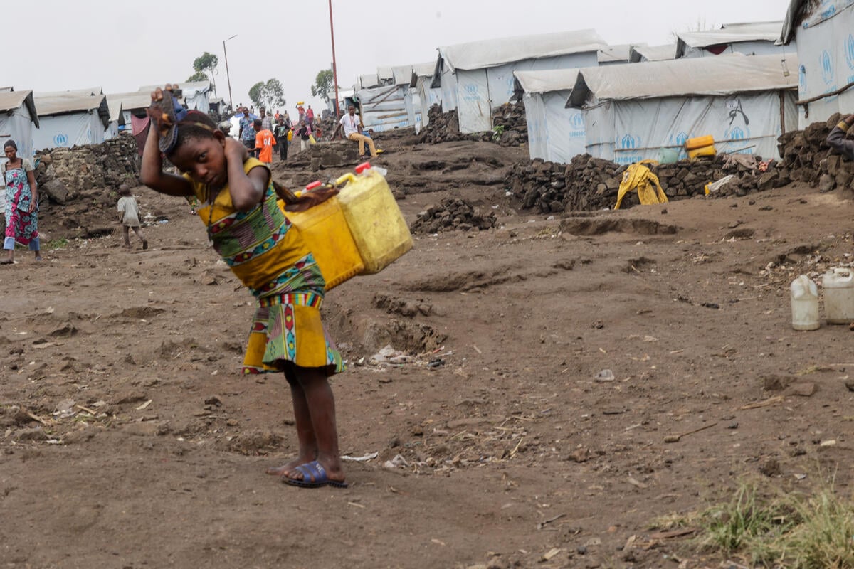 A small girl carries two yellow cannisters of water in a settlement for displaced people.