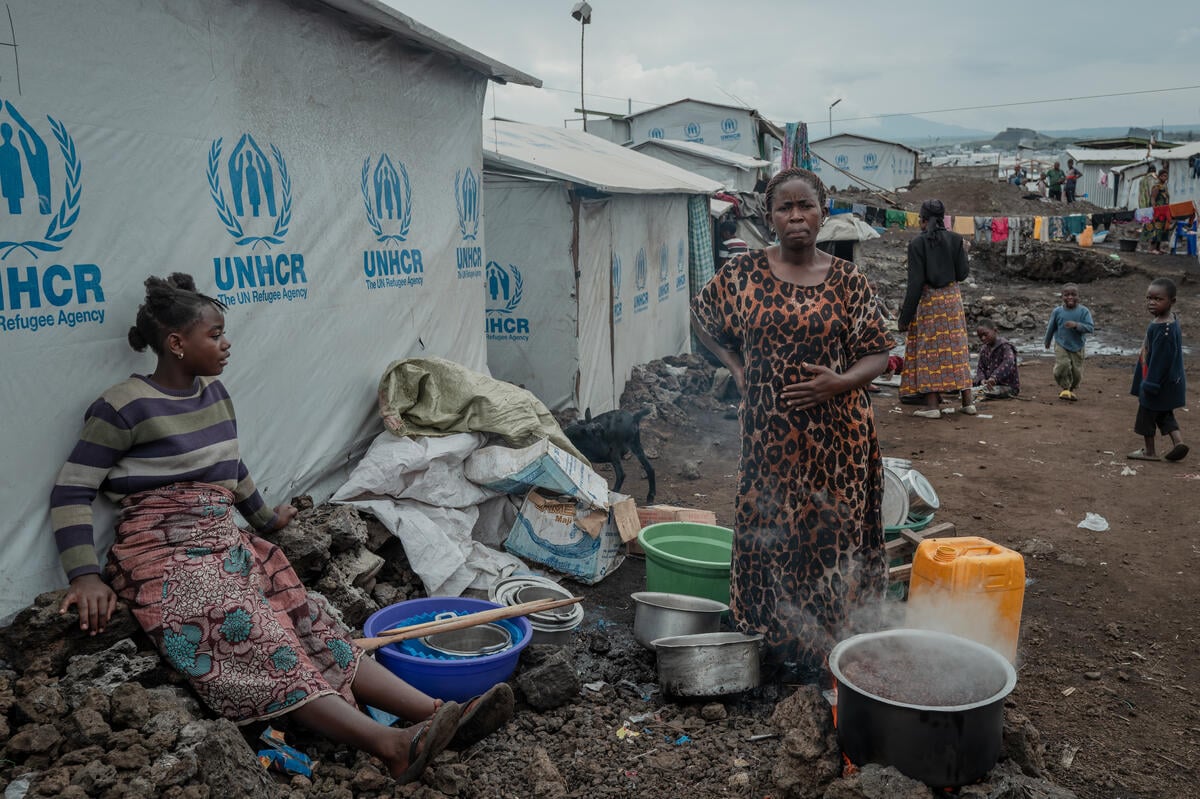 A woman stands behind a large pot cooking on an open fire outside a row of shelters with a girl sitting on a mound of stones behind the nearest shelter.