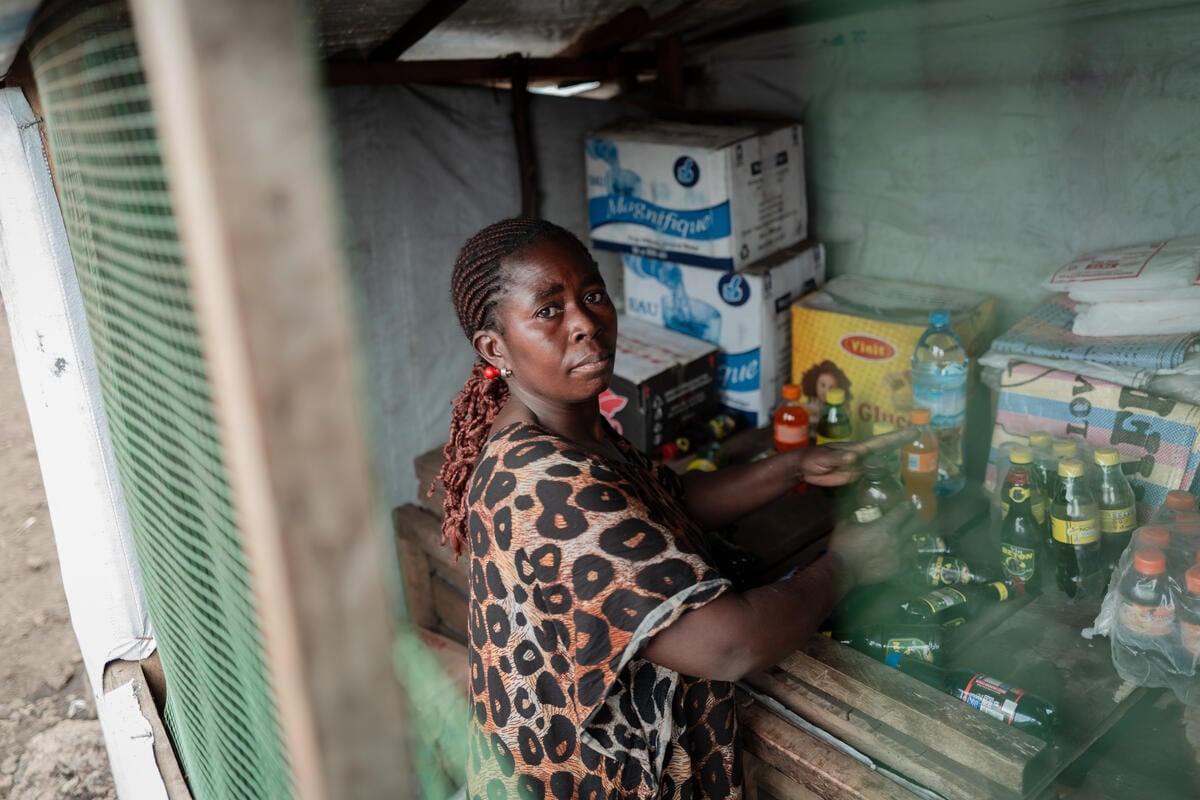 A woman arranges bottles on a wooden shelf inside a small store.