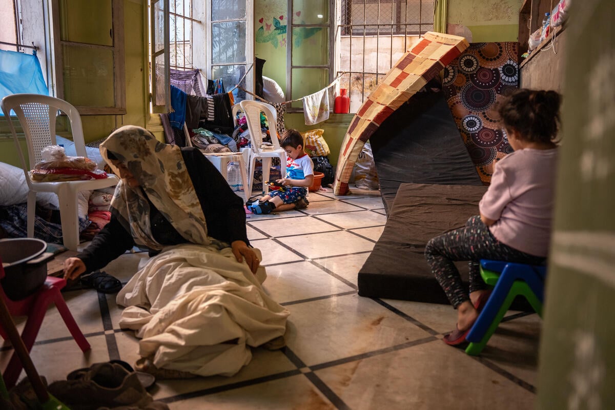 An elderly woman sits on the floor of a classroom filled with mattresses, clothes and chairs while two young children sit nearby.