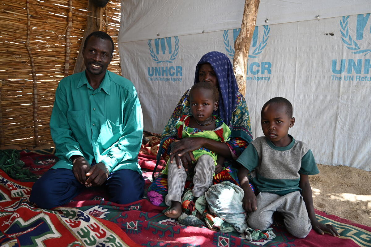 A couple sit on a carpet inside a woven reed shelter accompanied by two young children