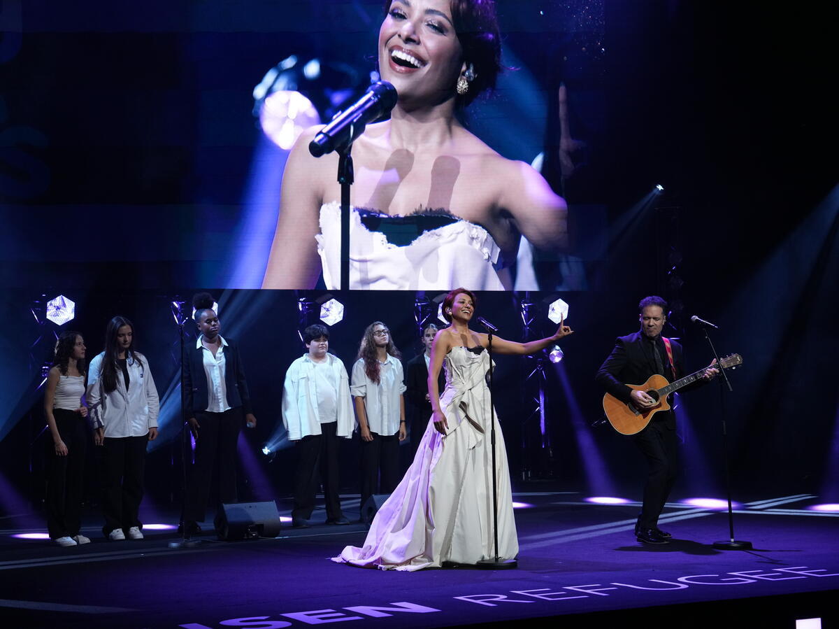 A woman performs onstage with a guitarist and backing singers in front of a giant screen