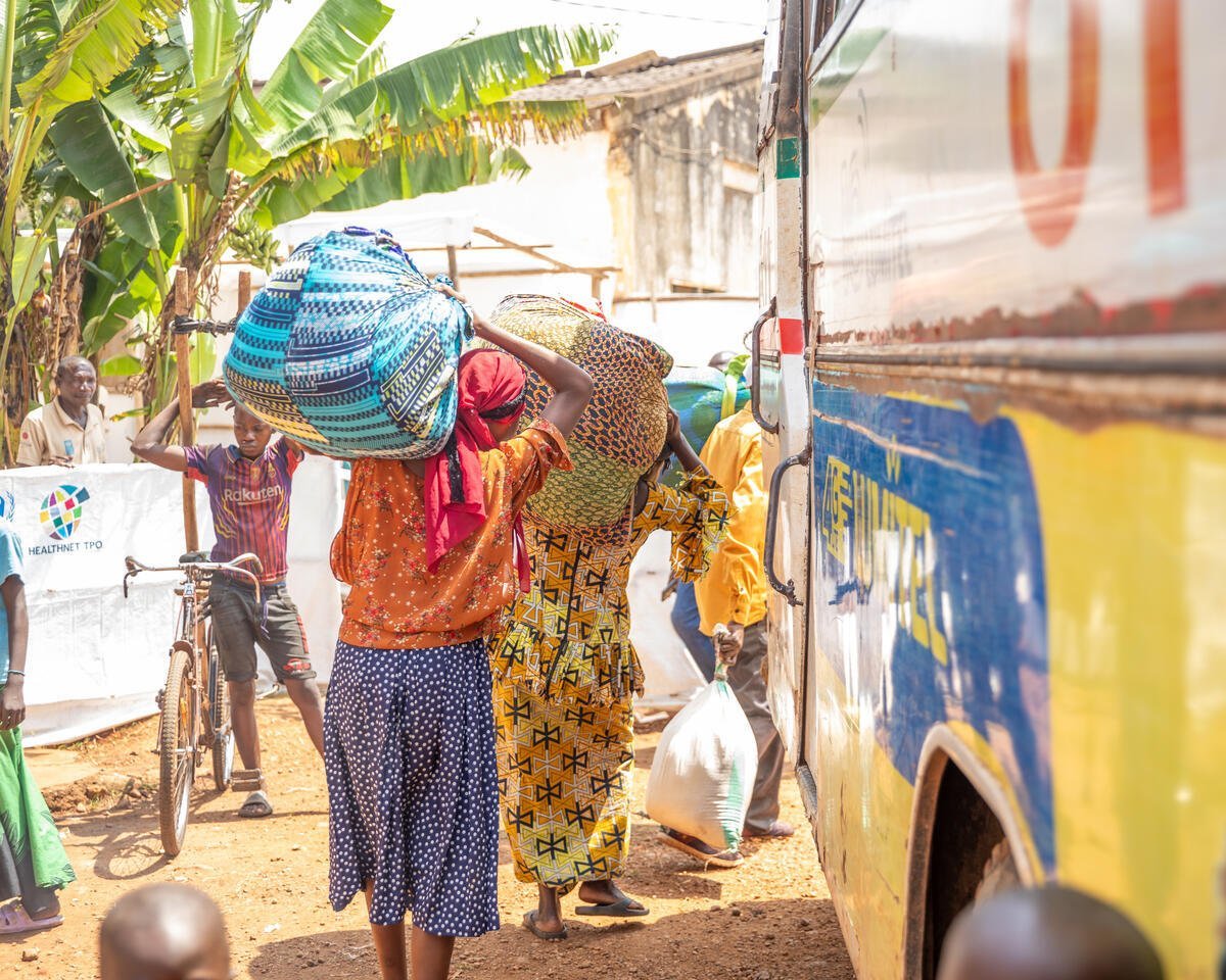 Three Congolese refugees load their belongings onto a bus while a young person stands nearby next to a bicycle, under a banana tree.