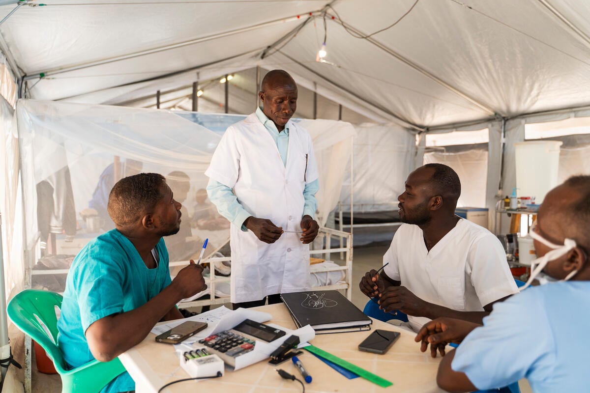 A male nurse in a white coat talks to his colleagues who sit at a round table in a hospital