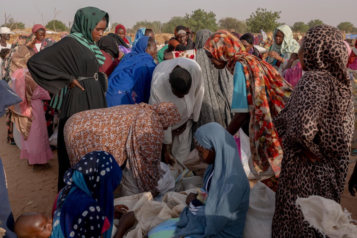 Outside in a sandy expanse with trees in the background, a large group of gather around sacks of grain on the floor