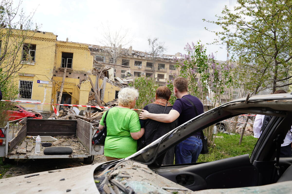 Three people pictured from behind, a damaged car behind them, as they observe a damaged building