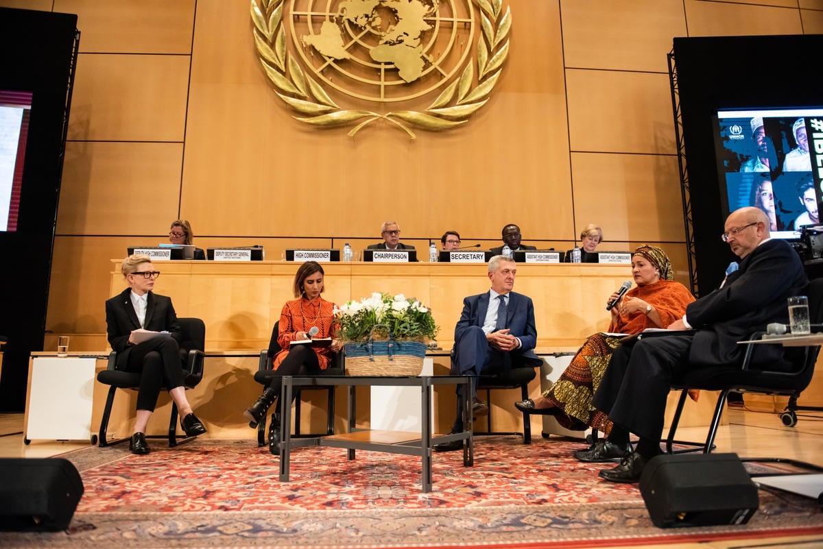 Four panellists and a moderator take part in a discussion, with several officials in the background.