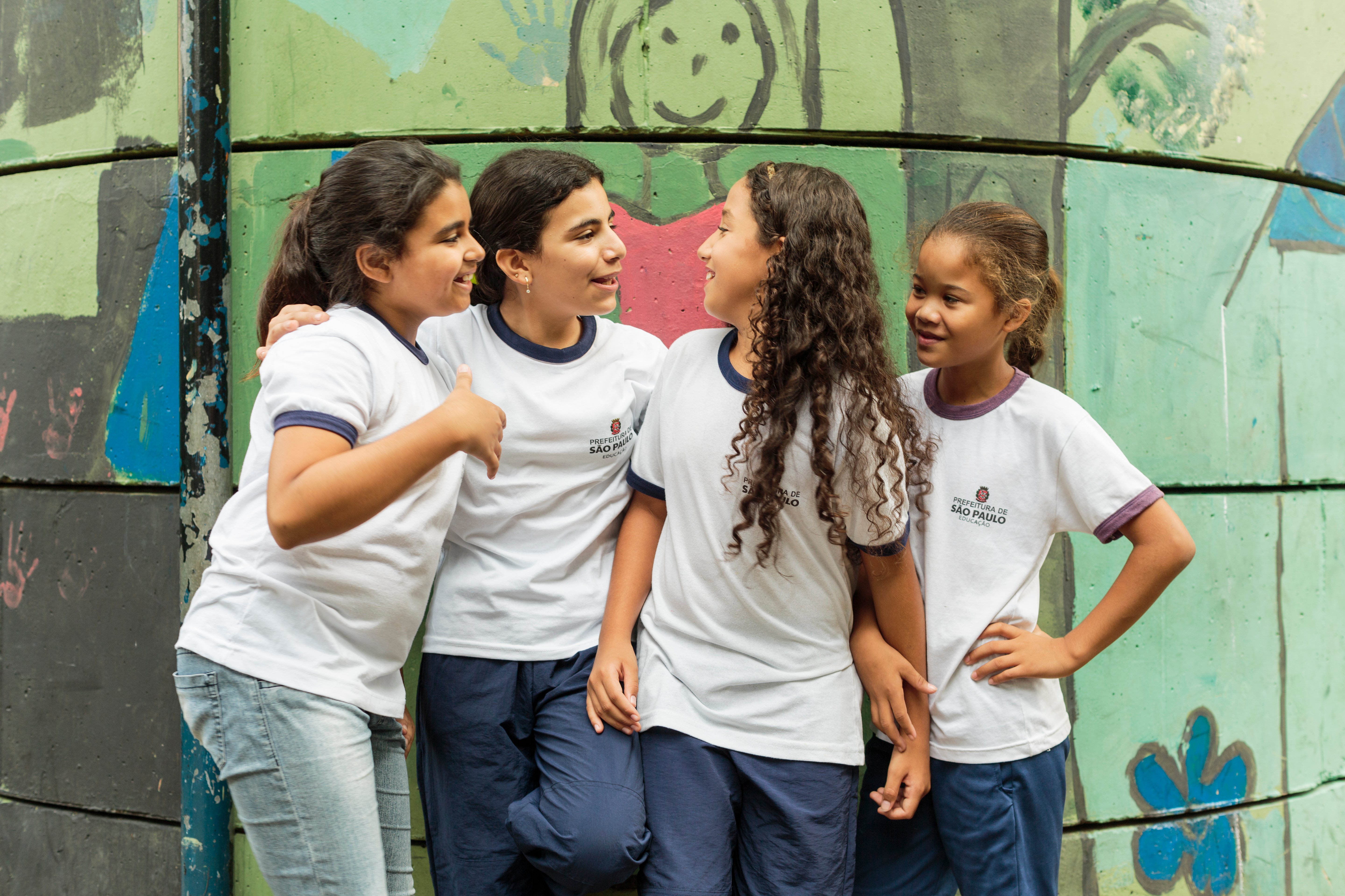 UNHCR UN Refugee Agency - How we do resettlement - ALT text: Four young girls enjoy chatting to each other while standing outside their classroom at the Duque de Caxias Municipal School in the Glicerio neighborhood of downtown Sao Paulo in Brazil.