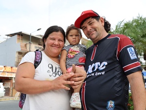 A man and a woman smile at the camera while holding their small child, buildings and a tree in the background.