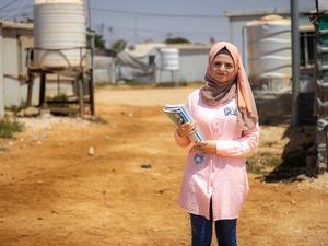 A young woman wearing a pink headscarf stands in a camp holding some school books.