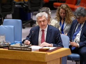 A man in a suit sits at a desk speaking into a microphone with other people seated behind him.