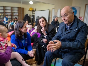 A man talks to a woman with a baby girl on her lap, seated in a communal room with other people in the background