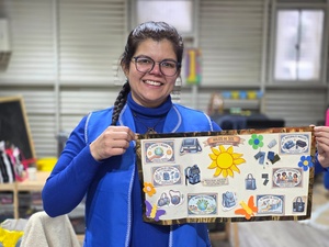 Una mujer con camiseta manga larga azul y chaleco azul, sostiene un tablero con recortes coloridos