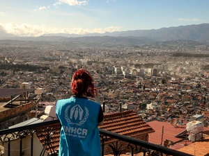 A woman in a UNHCR vest takes a photo of a city damaged by an earthquake.