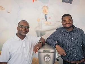 Two men stand either side of an industrial cake mixing machine in front of a mural-painted wall