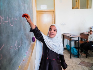An Afghan girl holds up her arm towards a blackboard in a classroom, as others sit at desks behind her.