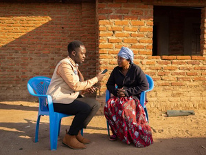 A man interviews a woman outside on two plastic chairs, in front of a brick building