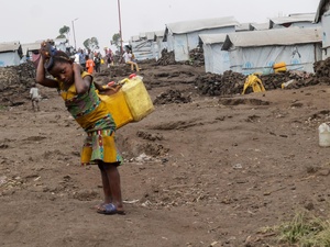 A young girl with two jerry cans on her back stands in front of a row of tents covered in tarpaulin.