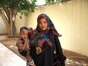 A woman wearing a headscarf stands in a walled courtyard holding a toddler.