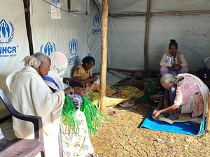 Several women sit inside a UNHCR tent and craft woven items