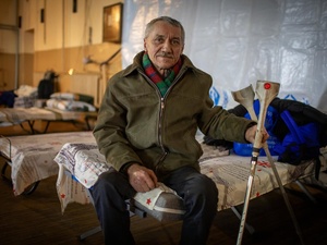 An older man sits on a camp bed in a dormitory, holding some crutches.