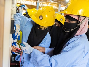 Two women wearing niqabs and yellow hardhats work together to screw a switch to a wall while other women look on.