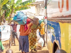 Three Congolese refugees load their belongings onto a bus while a young person stands nearby next to a bicycle, under a banana tree.