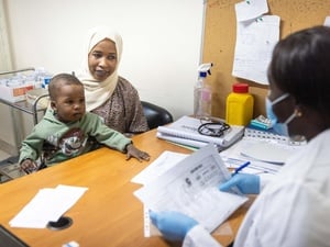 A woman is seated at a table with her young son, across from a medical staff member staff in a mask who is looking at paperwork
