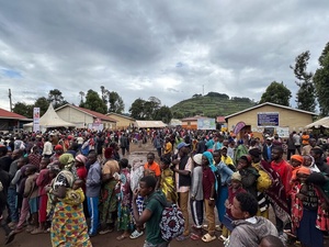 Large groups of Congolese refugees gather outside some buildings and shelters with trees and hills behind them.