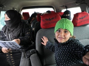 A young girl wearing a knitted green hat sits inside a minibus with women passengers sitting in the seats behind her.