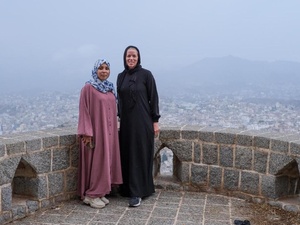 Two women stand on a stone terrace with a city landscape behind them