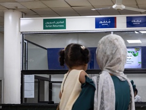 A woman carrying her daughter stands in front of a reception counter with a sign reading 'Syrian returnees' in Arabic and English