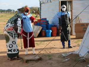 Health workers wearing face masks are pictured outdoors with disinfection equipment. They are standing in front of a metal shed and piles of buckets.