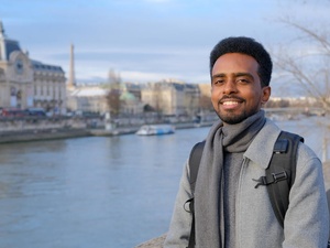 A man wearing a grey overcoat and backpack stands on a riverside with historic buildings on the opposite bank 