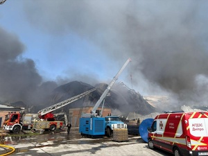 Black smoke surrounds a damaged warehouse, with emergency vehicles attending