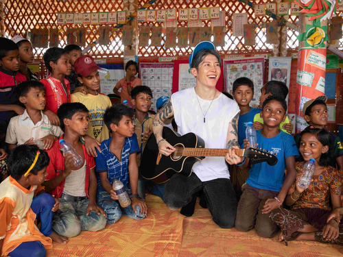 MIYAVI kneeling on the floor with his guitar, smiling, a group of children around him