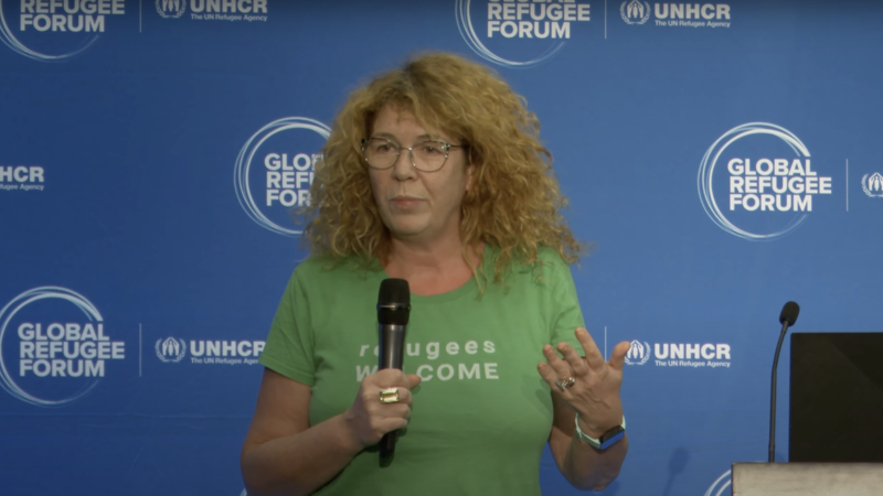 A woman in a green t-shirt stands in front of a blue sign holding a mic