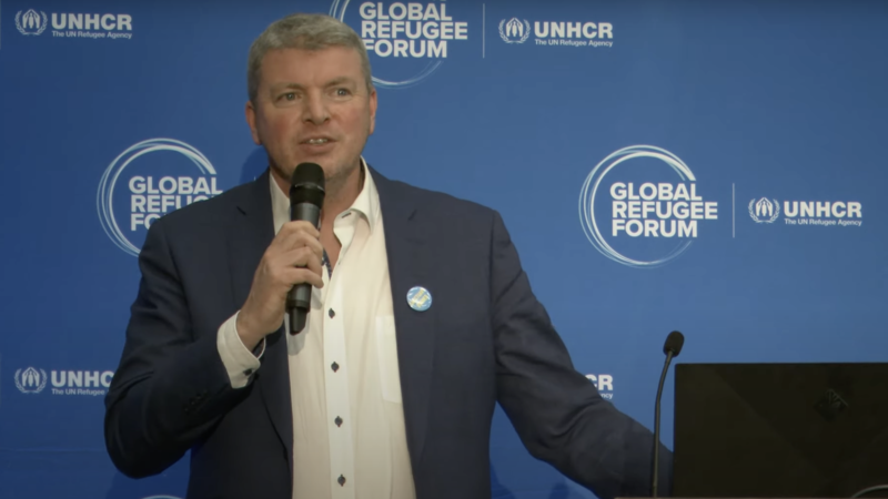 A man in a suit holds a mic in front of a blue wall.