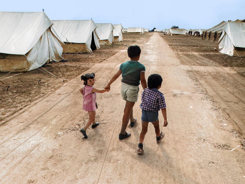 Three children walk through a refugee camp of white tents. The photo originally was originally shot in black and white and has been colourized. 