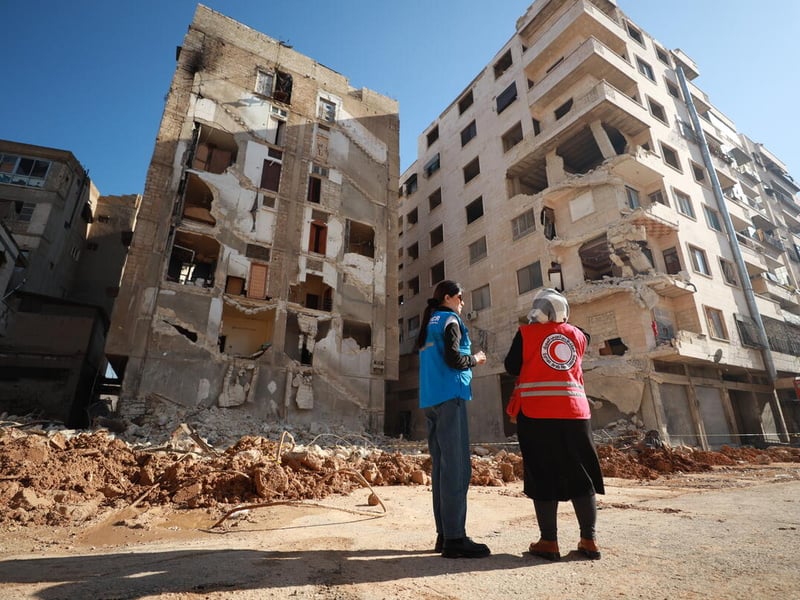 A UNHCR staff member and Red Cross staff member stand in front of a destroyed building.
