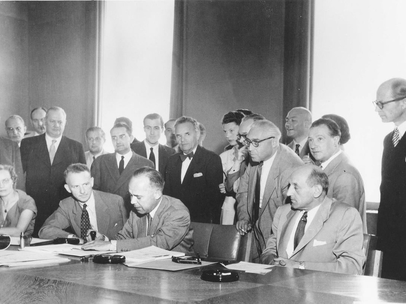 Back and white photo: A man signs the 1951 Refugee Convention, surrounded by a group of people.