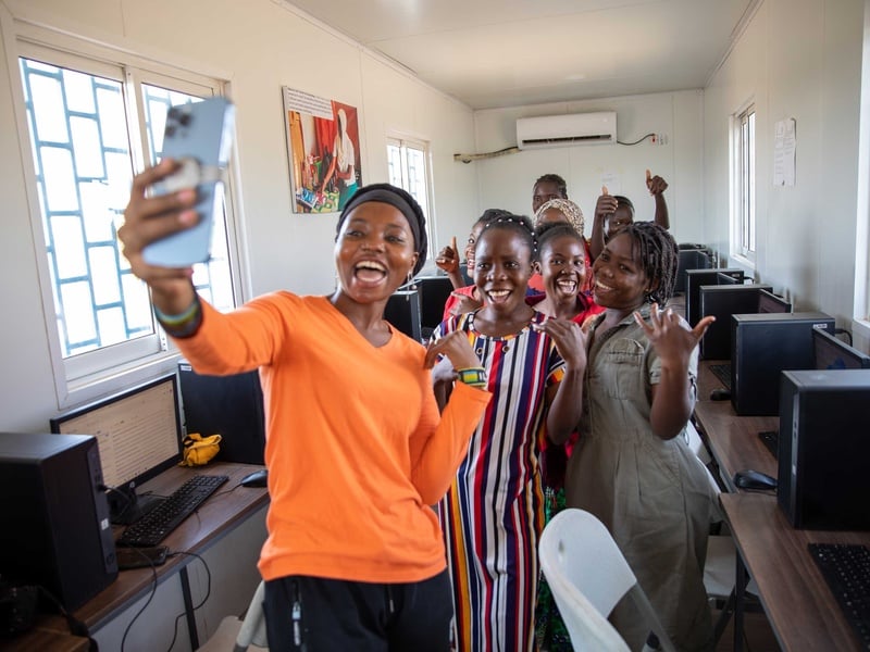 A woman in a bright orange shirt takes a selfie with a group of young women in a computer lab.