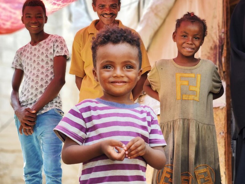 A three-year-old internally displaced boy from Yemen smiles for the camera. His father and older siblings are standing behind him.