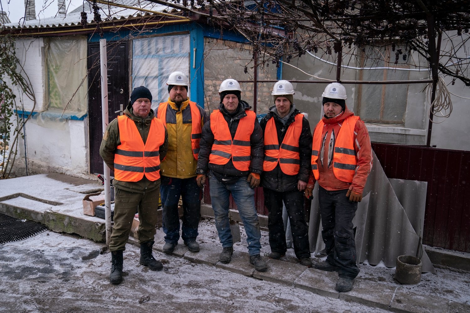 A group of contruction workers wearing high-vis vests stands outside a partially damaged house in Ukraine.