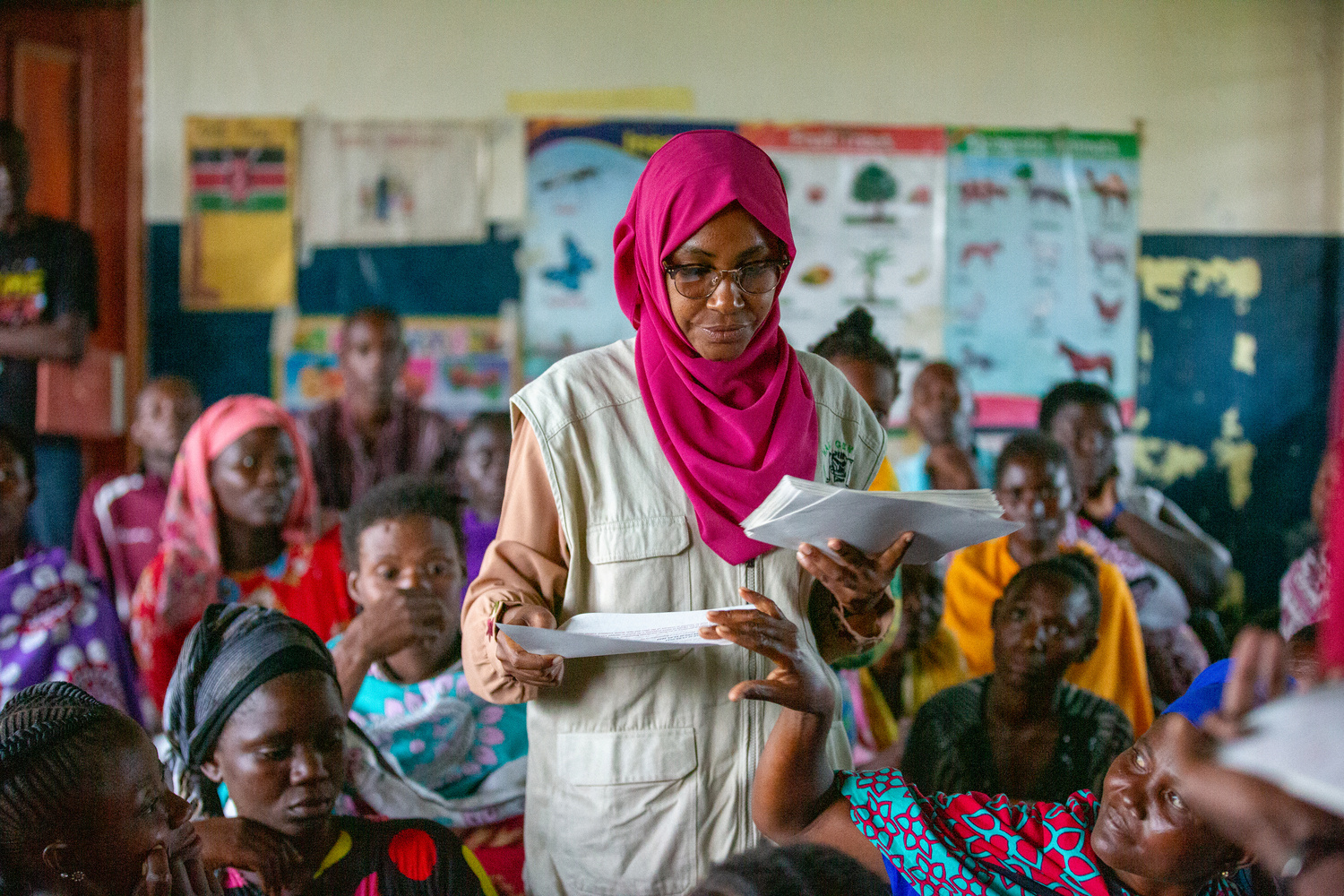A woman in a pink headscarf stands in a classroom handing out papers to other women who are seated.