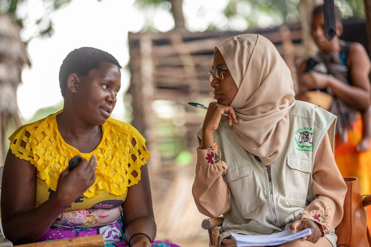 A woman in a headscarf holding some papers and a pen listens to a woman seated next to her in a yellow top.