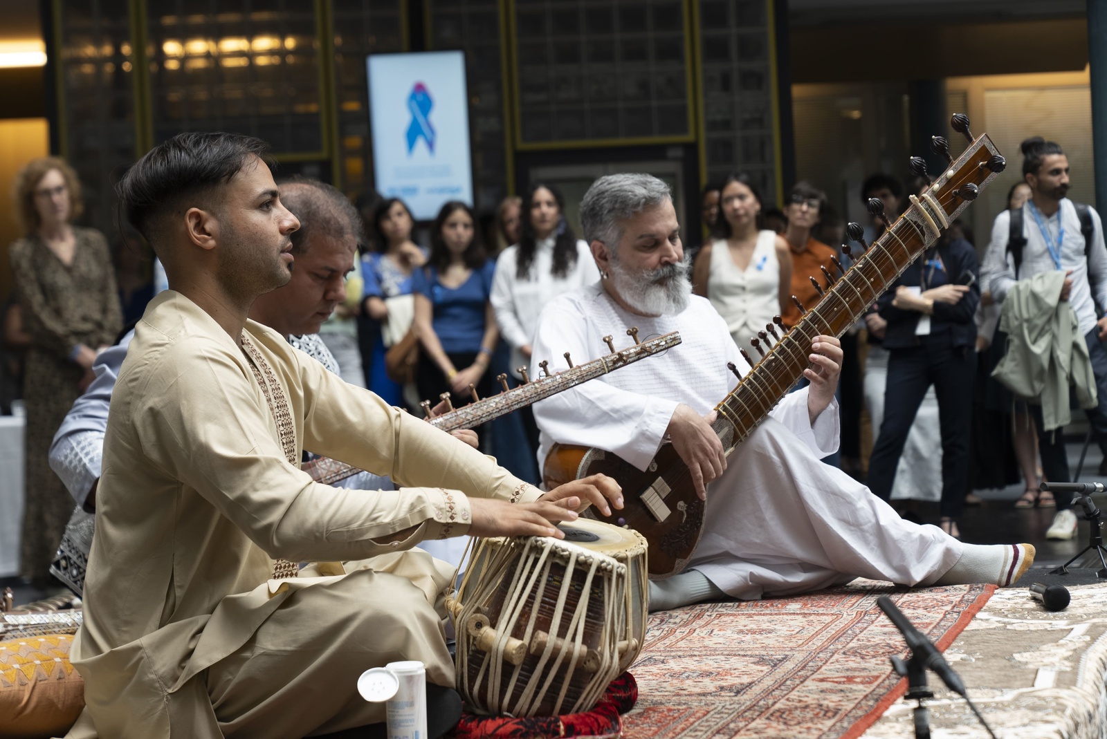 Three men wearing traditional dress and seated on a rug play instruments in the lobby of an office building