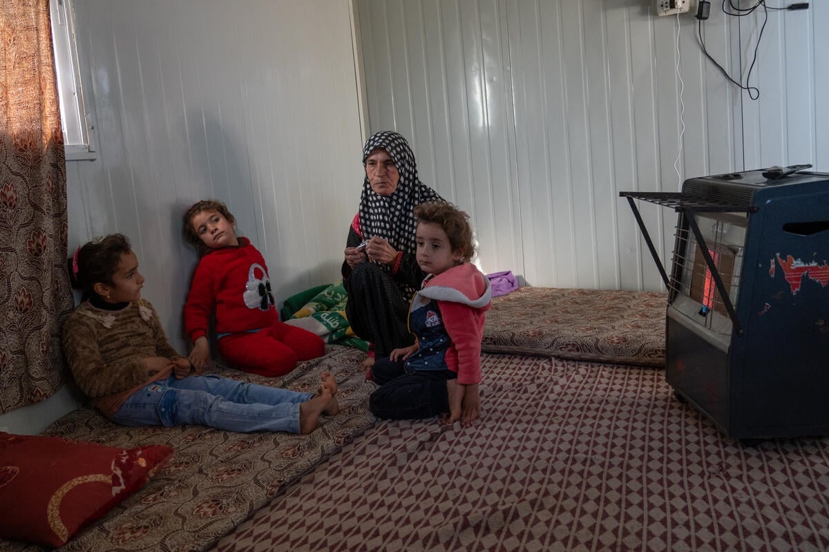 A woman sits with her grandchildren in front of a gas heater inside her shelter.