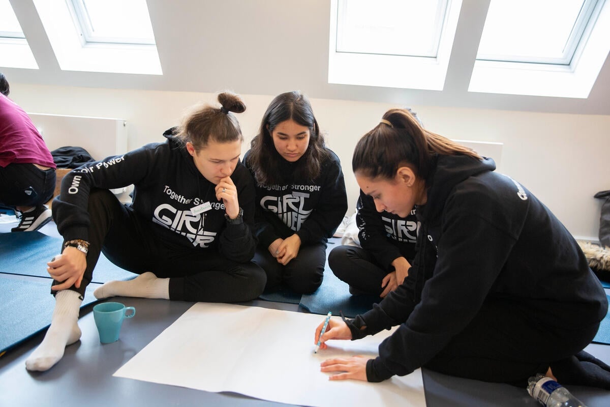 Four women sit on the floor, gathered around a large piece of paper