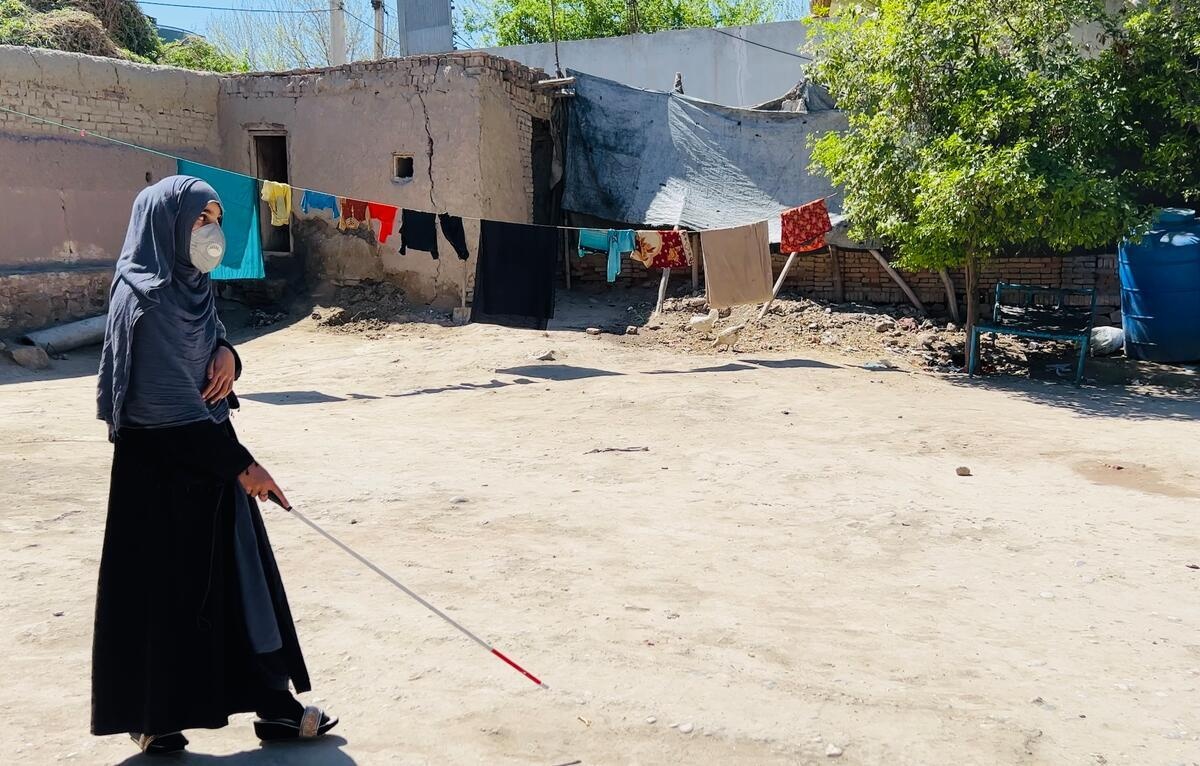 Amina walks along a dirt path with the help of a guide cane.